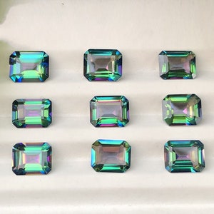 May include: Nine emerald green, iridescent, rectangular gemstones with a faceted cut. The gemstones are arranged in three rows of three on a white surface.