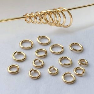 Könnte beinhalten: Eine Sammlung goldfarbener Biegeringe in verschiedenen Größen, einige offen und einige geschlossen, auf einem goldenen Stab dargestellt. Die Ringe sind auf einer weißen Oberfläche verstreut und zeigen ihre kreisförmige Form und ihren metallischen Glanz. Ideal für die Schmuckherstellung.