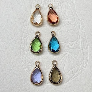 May include: Six teardrop-shaped pendants with gold-coloured settings. The pendants feature faceted gemstones in clear, peach, green, blue, purple, and brown hues. Each pendant has a small loop at the top for attaching to a chain or other jewellery component.