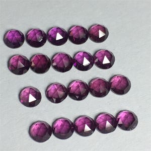 May include: A collection of 17 round, faceted, purple gemstones. The gemstones are arranged in rows on a light grey surface.