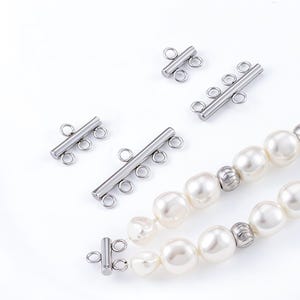 May include: Close-up of jewelry components, including several silver-colored bar clasps with multiple loops and a strand of large, off-white pearls. The clasps are designed for multi-strand necklaces or bracelets.