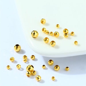 May include: A collection of gold-colored metal beads with a faceted design. The beads are scattered on a white surface.