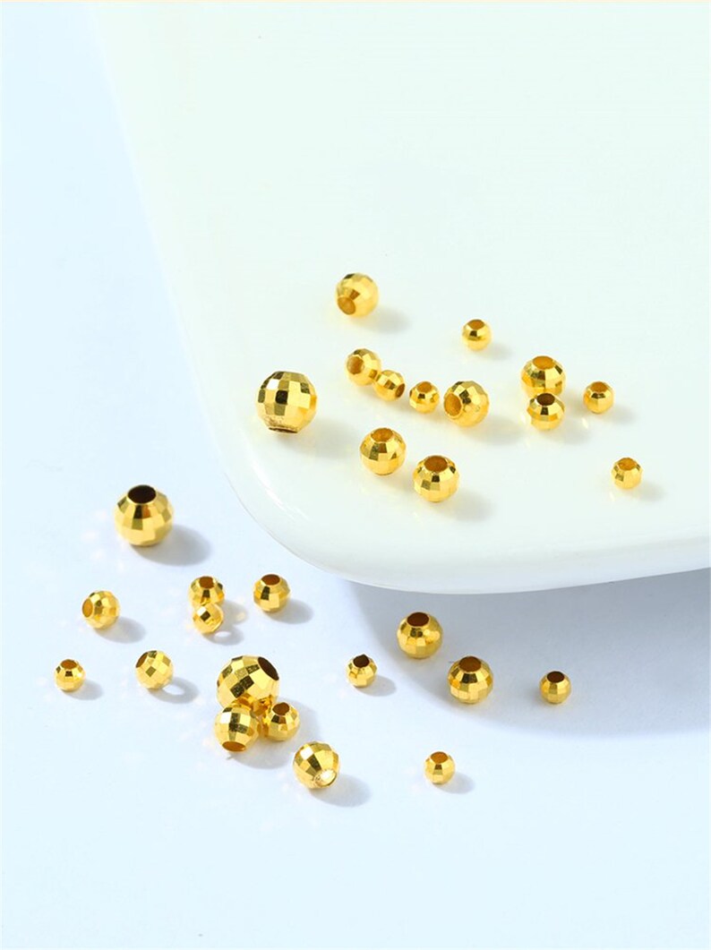 May include: A collection of gold-colored metal beads with a faceted design. The beads are scattered on a white surface.