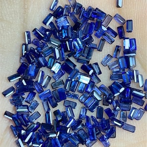 Puede incluir: Una colección de pequeñas piedras preciosas de zafiro azul rectangulares. Las piedras preciosas son facetadas y tienen una variedad de tamaños y formas.