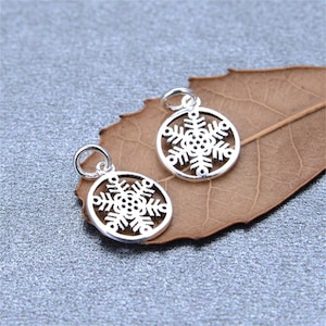 May include: Two silver-toned metal charms, each featuring a circular frame enclosing a detailed snowflake design. Each charm includes a small jump ring at the top for attachment, showcasing a polished finish and intricate cut-out patterns.