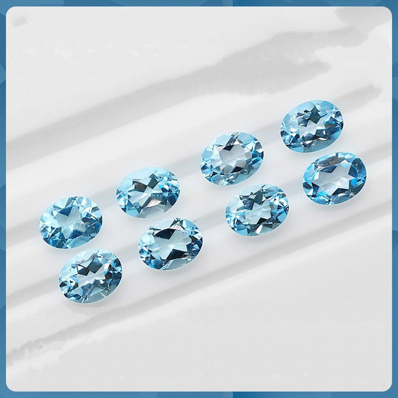 May include: Eight oval-shaped, light blue gemstones on a white surface. The gemstones are arranged in a semi-circle.
