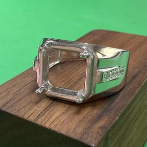 Op de afbeelding: Een zilveren ring met een vierkante zetting voor een edelsteen. De ring heeft een band met kleine, heldere stenen die erin zijn gezet.
