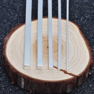 May include: Five silver-coloured metal square rods of varying widths arranged vertically in a row. The rods have a smooth, reflective finish and a consistent square cross-section, displayed side-by-side to show the clear difference in their thickness.