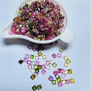 May include: A white, scoop-shaped dish overflowing with a collection of small, square-cut gemstones. The gemstones display a variety of colors, including pink, green, yellow, and purple. Some gemstones are scattered on the white surface.