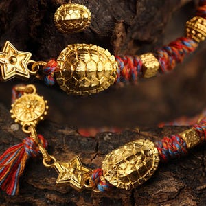 May include: Gold-colored bracelets with turtle and star charms. The bracelets feature red, blue, and yellow braided cords. One bracelet has a tassel. The charms have a textured surface. The background is a dark, textured surface.