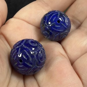 Könnte beinhalten: Zwei runde, blaue, geschnitzte Lapislazuli-Perlen. Die Perlen haben ein detailliertes, in die Oberfläche geschnitztes Blumenmuster.