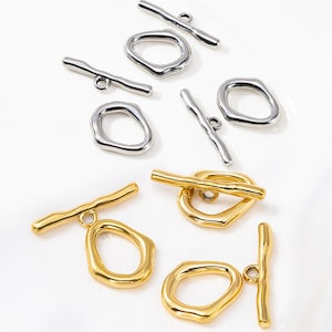 Puede incluir: Una variedad de cierres de joyería en tonos plateados y dorados. Los cierres presentan un diseño de barra y anillo, con una forma irregular y orgánica. El fondo es una superficie blanca lisa, que resalta los hallazgos de joyería.