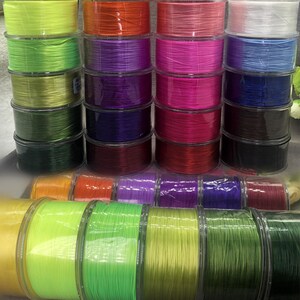 Könnte beinhalten: Eine Sammlung von Angelschnurspulen in verschiedenen Farben, darunter Grün, Orange, Lila, Pink und Blau. Die Spulen sind in Reihen gestapelt und zeigen die verschiedenen Farbtöne der Angelschnur.