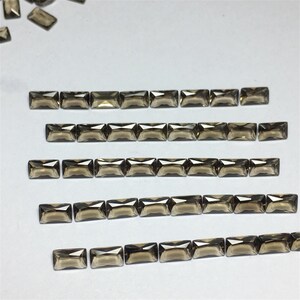 May include: Several rows of rectangular, smoky-brown gemstones are displayed on a white surface. The gemstones are faceted, catching the light. The arrangement suggests a collection of loose stones, possibly for jewellery making or other crafts.
