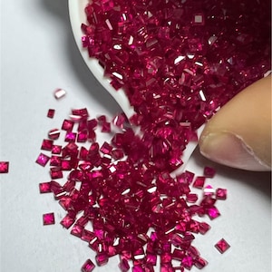 Könnte beinhalten: Nahaufnahme eines Haufens kleiner, quadratischer, roter Edelsteine. Die Edelsteine liegen lose und verstreut auf einer weißen Oberfläche.