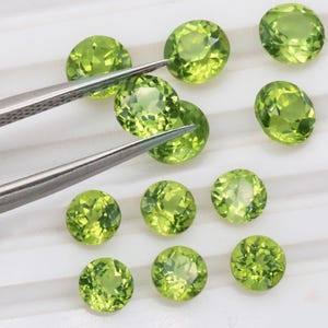 Puede incluir: Un conjunto de gemas de peridoto redondas y facetadas de varios tamaños. Las gemas son de un color verde claro vibrante y se muestran sobre una superficie blanca. Unas pinzas sostienen una de las piedras, resaltando su claridad.