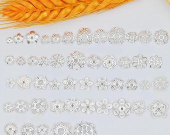 May include: An assortment of silver-colored, flower-shaped bead caps in various sizes and designs. The caps have intricate cut-out patterns and are arranged on a white surface with a golden wheat stalk in the background.