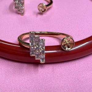 May include: A gold ring with a row of clear cubic zirconia stones set in a prong setting. The ring is open on one side and has a round bezel setting on the other side.