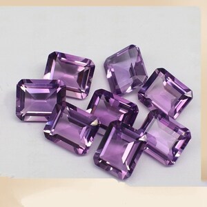 May include: A group of eight purple amethyst gemstones, cut in a rectangular emerald shape. The gemstones are faceted and have a shiny, polished finish.