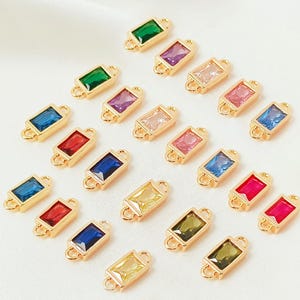 May include: A selection of gold-coloured rectangular connectors, each featuring a rectangular gemstone in various colours. The gemstones include green, purple, pink, blue, red, and yellow. Each connector has loops for attachment to other items.
