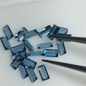 May include: A collection of loose blue topaz gemstones in a rectangular cut. The gemstones are arranged on a white surface and are being held by tweezers.