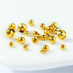 May include: A collection of gold-colored metal beads with a faceted design. The beads are scattered on a white surface.