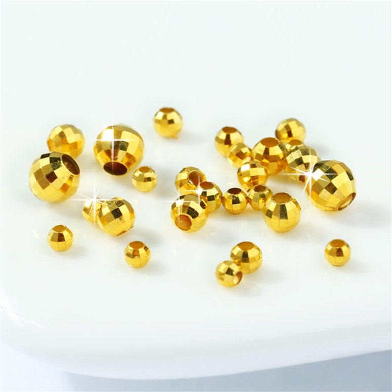 May include: A collection of gold-colored metal beads with a faceted design. The beads are scattered on a white surface.