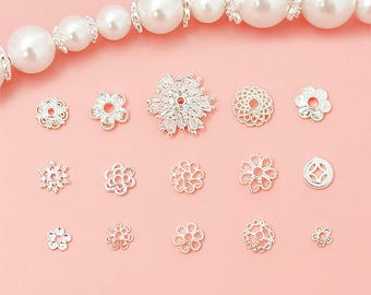 May include: An assortment of silver-toned jewelry findings, including flower-shaped beads and decorative accents, are displayed against a soft pink background. A strand of large, white faux pearls is at the top. These findings are ideal for jewelry making.