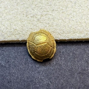 May include: A small, gold-colored turtle shell charm. The charm has a textured surface with a detailed pattern of segmented plates. The charm is likely made of metal and is displayed on a dark surface.