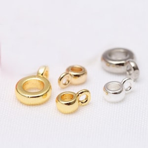 May include: Close-up of several small jewelry findings in gold and silver tones. The findings have a circular design with a small loop attached, perfect for jewelry making. The background is a soft white fabric.