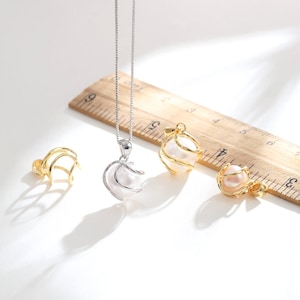 May include: A silver-toned necklace with a pearl pendant encased in a wire cage, accompanied by three matching gold-toned cage charms. A wooden ruler is visible, showing the charms measure approximately 0.5 inches in diameter.