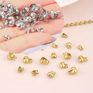 May include: A collection of silver and gold-colored ball chain connectors. The silver connectors are held in a hand, while the gold connectors are scattered on a pink surface. The connectors are small, with a rounded ball shape and a clasp.