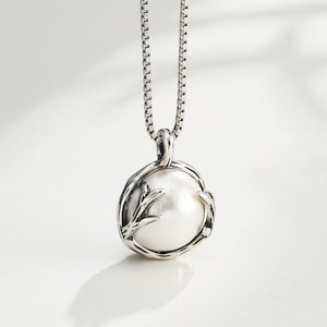 May include: A sterling silver pendant necklace featuring a round, lustrous white pearl encased in a textured, branch-like metal setting. The pendant hangs from a delicate box chain, showcasing oxidized silver details that contrast with the pearl.