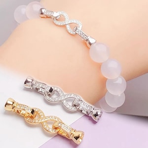 May include: A beaded bracelet with large, pale pink beads and a silver-colored infinity clasp. Two additional clasps, one silver and one gold, are displayed below. The clasps are adorned with small, clear stones.