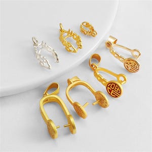 May include: A collection of metal jewelry bails in gold and silver tones. These U-shaped findings feature circular side accents with embossed patterns, including one with a red circular inlay. Each piece includes a top loop for attachment.