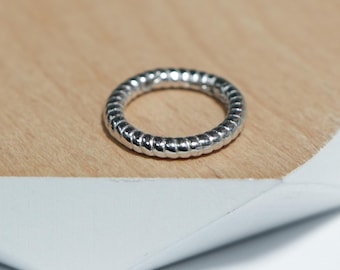May include: A silver ring with a textured, ridged design. The ring is circular and sits on a light brown surface. The ring's texture creates a pattern of small, repeating lines around its circumference.