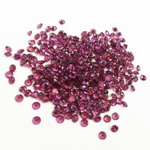 May include: A large pile of small, round, purple gemstones. The gemstones are scattered on a white surface.