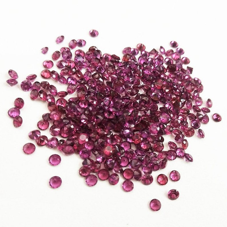 May include: A large pile of small, round, purple gemstones. The gemstones are scattered on a white surface.