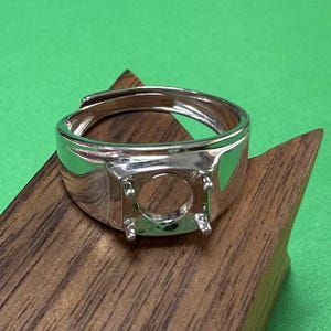 May include: A silver ring with a round bezel setting for a gemstone. The ring is on a wooden surface.
