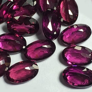 Puede incluir: Un surtido de gemas facetadas de forma ovalada en varios tamaños. Las gemas son de un color magenta intenso, con múltiples facetas que capturan la luz. Las piedras preciosas están dispersas sobre una superficie blanca.