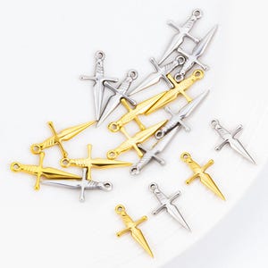 Peut inclure: Un assortiment de petits charms en forme de dagues, de couleur or et argent. Les charms ont une garde détaillée et une lame pointue. Les dagues sont disposées sur une surface blanche. Idéal pour la création de bijoux ou les projets d'artisanat.