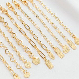 May include: A collection of gold-colored chain extenders with various link styles, including oval, heart, and round designs. Some chains feature small charm pendants with engraved details. The extenders have lobster claw clasps.