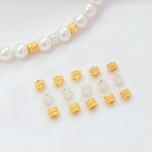 May include: An assortment of gold and silver-colored decorative beads. Some beads are incorporated into a pearl necklace. The beads have a detailed, textured design. The background is a white, textured surface.