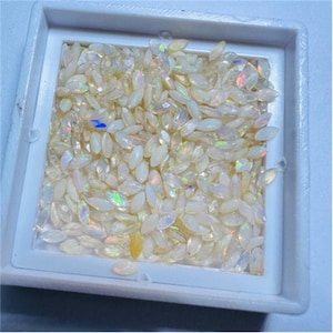 May include: A close-up view of a white, oval-shaped opal gemstones. The opals are loose and scattered in a white tray. The opals have a slight iridescent sheen.