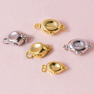 Può includere: Cinque chiusure per gioielli in tonalità oro e argento sono disposte su una superficie rosa chiaro. Le chiusure sono di forma ovale con una rientranza centrale e piccoli anelli su ogni lato per attaccare i gioielli. Le chiusure sono progettate per fissare collane o bracciali.
