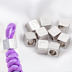 Puede incluir: Cubos plateados con letras del alfabeto y un cordón trenzado morado y blanco. Los cubos están grabados con letras. El cordón tiene dos piezas cuadradas plateadas con las letras "XX".