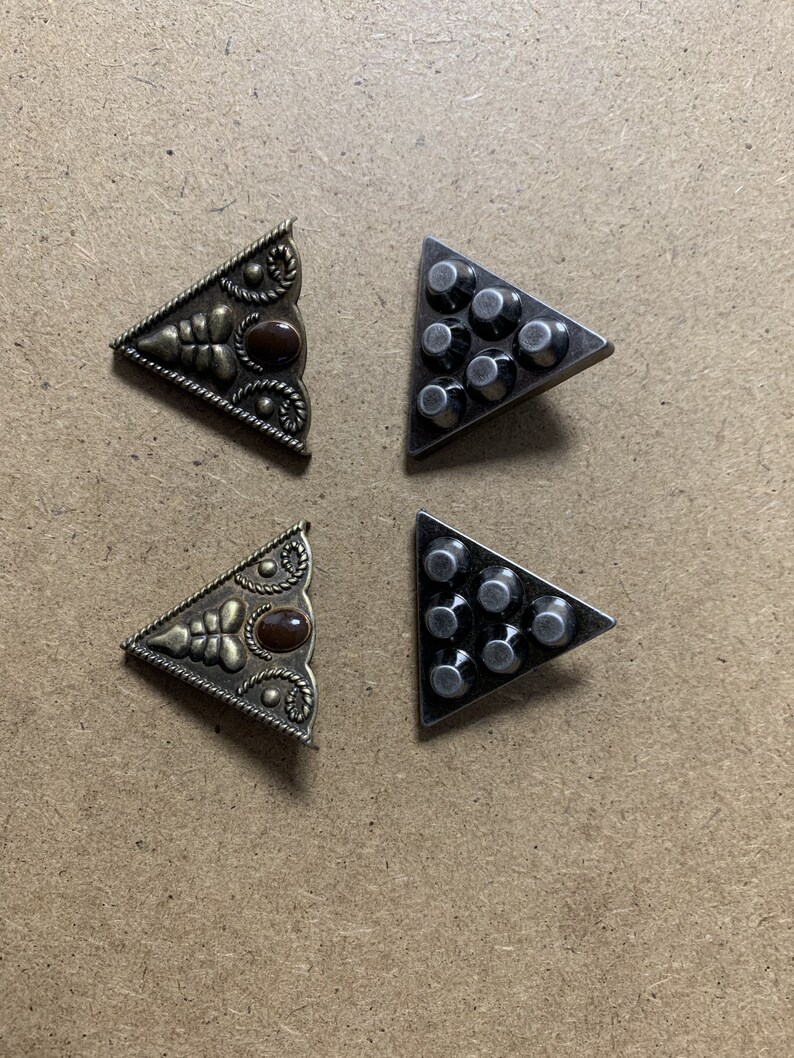 May include: Four triangle-shaped metal embellishments. Two are ornate with a brown stone and gold-toned metal. Two are plain with a silver-toned metal and a series of raised dots.
