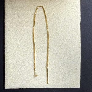 Könnte beinhalten: Ein goldener Kettenohrring mit einem langen, dünnen, geraden Stück Metall, das von unten hängt.