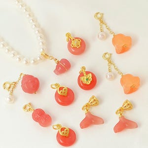 May include: A collection of gold-toned pendants and charms in various shapes, including round, flower, and peanut designs. Some charms feature pearls and a butterfly. The pendants are in shades of pink and orange, with a white pearl necklace.
