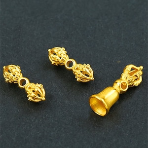 May include: Three gold-colored metal charms in the shape of a bell and two double-sided prayer wheels. The prayer wheels have intricate designs and are connected by a small ring.